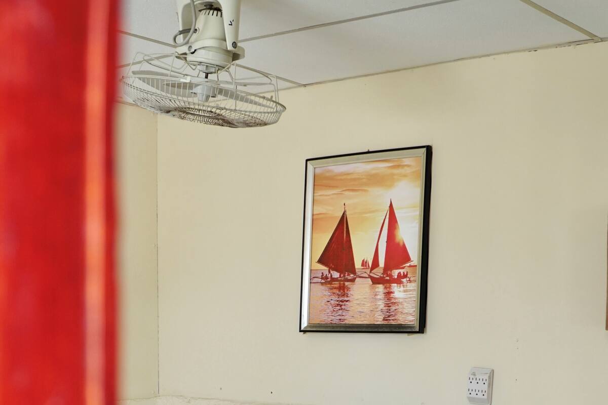 Uma cama de solteiro com colchão e travesseiros brancos, um criado-mudo de madeira com abajur, um ventilador de teto e um quadro com barcos à vela na parede.