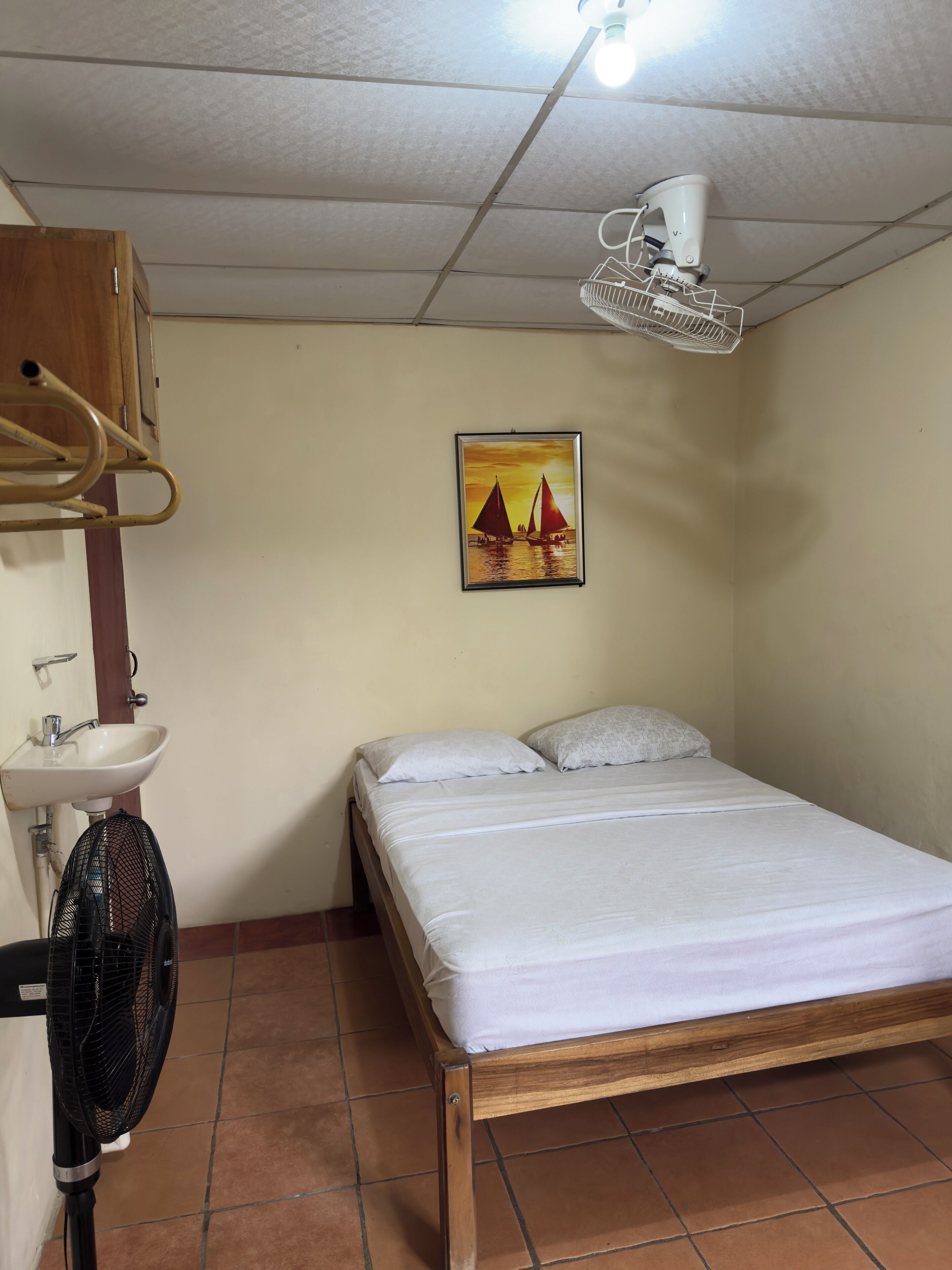 Un lit simple avec un cadre en bois, un matelas blanc et deux oreillers. Un ventilateur de plafond blanc est fixé au mur, et un tableau représentant des voiliers est accroché au-dessus du lit.