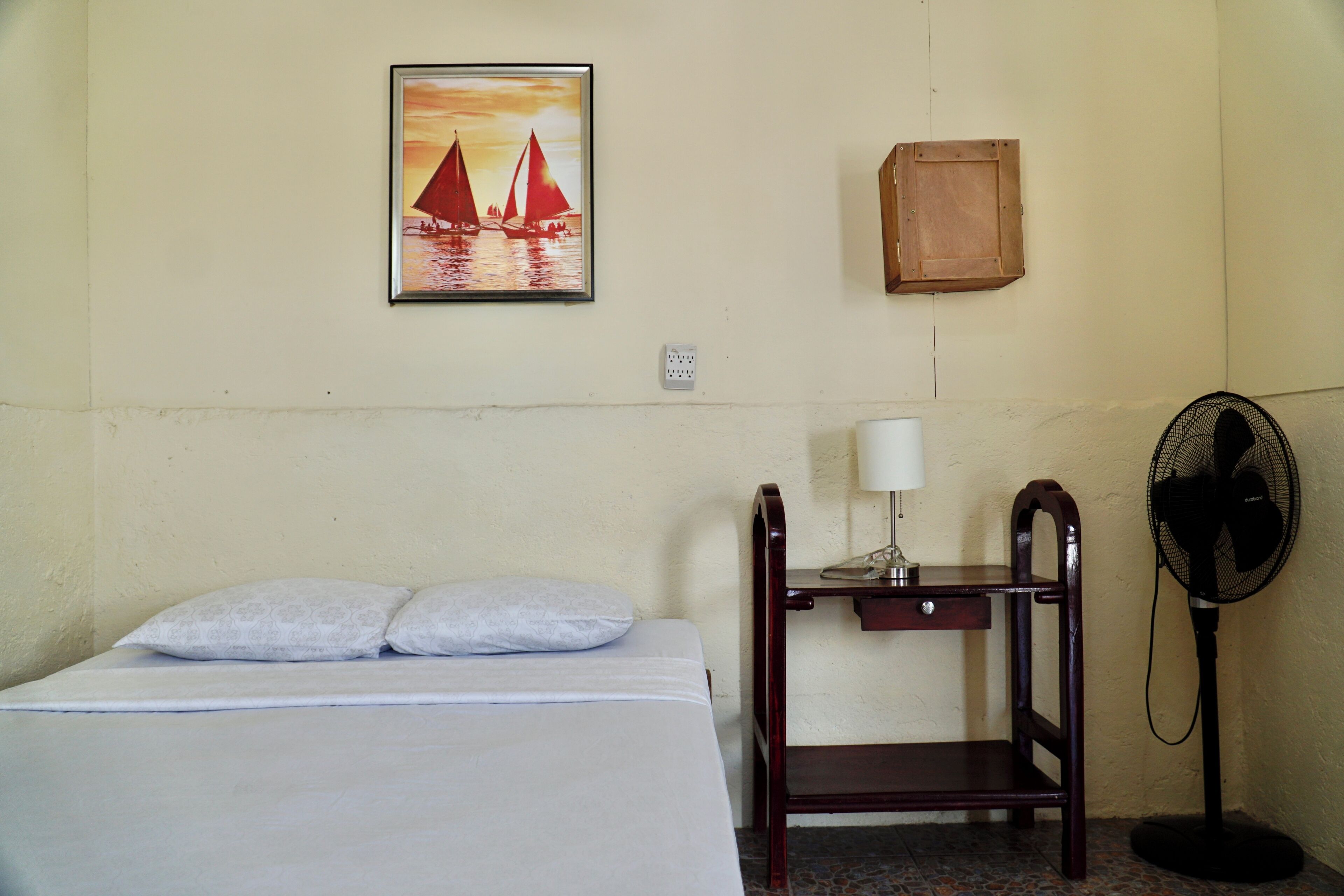 Une chambre d’hôtel avec un lit, un bureau doté d’une lampe, un ventilateur, un meuble fixé au mur et un tableau représentant des voiliers.
