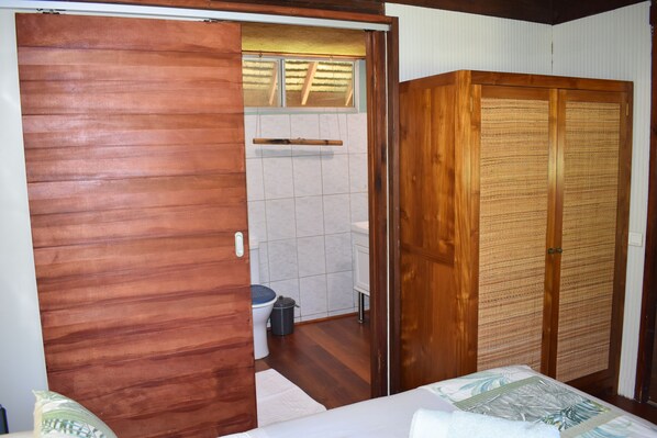 Un armario de madera con puerta trenzada, una cama con cubrecama blanco y un baño pequeño con inodoro y cubo de basura.