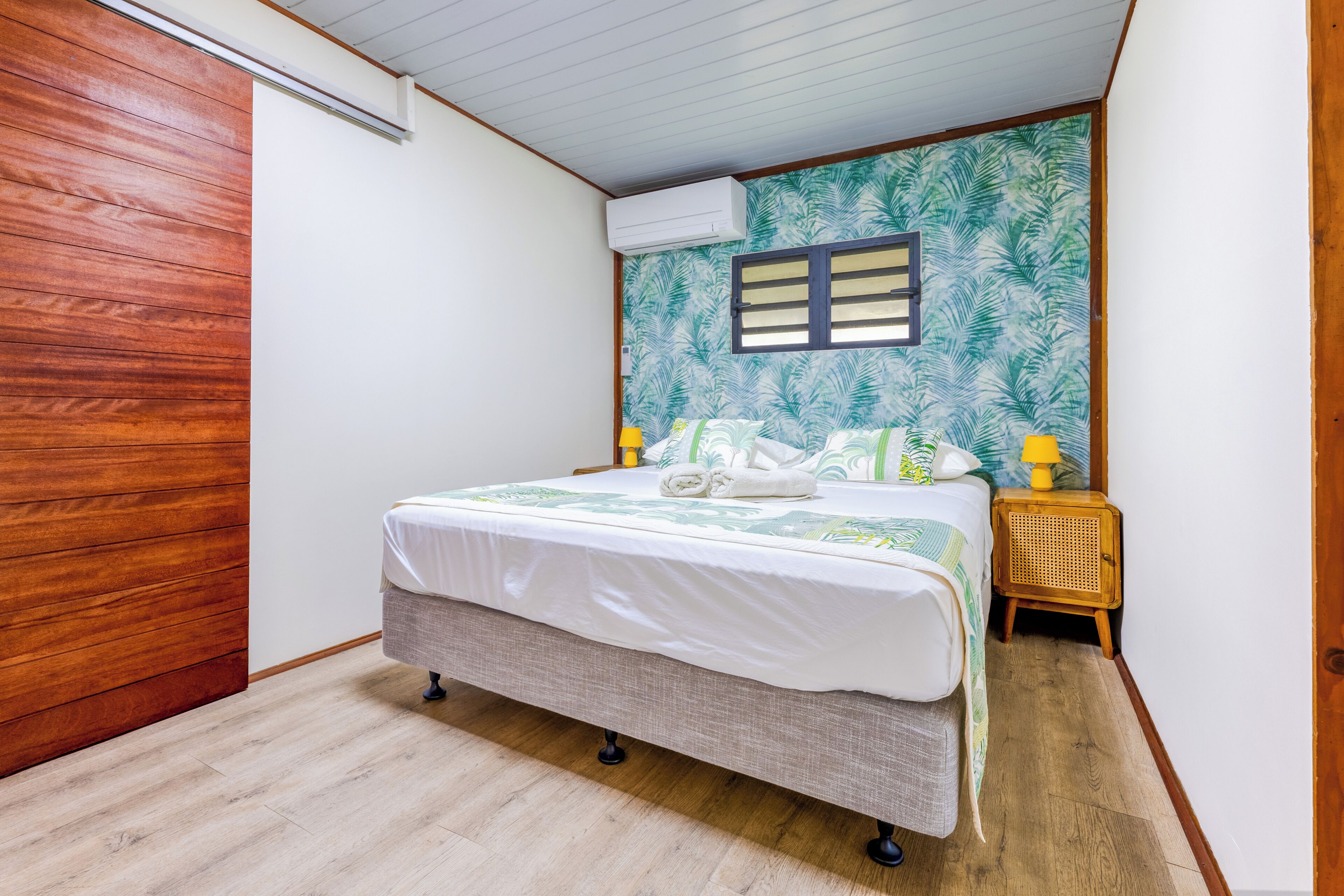 Un dormitorio con cama, una puerta de madera, aire acondicionado instalado en la pared, una ventana con persianas y una mesita de noche con lámpara.