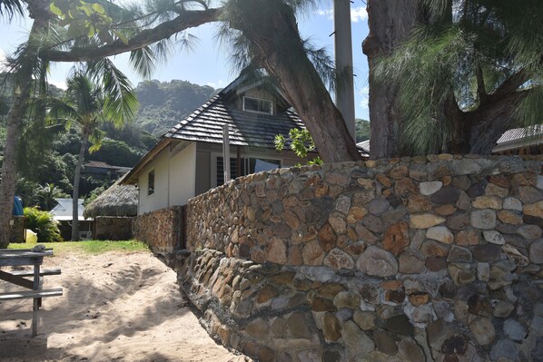 A house with a stone wall, a thatched-roof structure, and a sandy area with a picnic table. - Fare Aute Beach (Moorea-Maiao)
