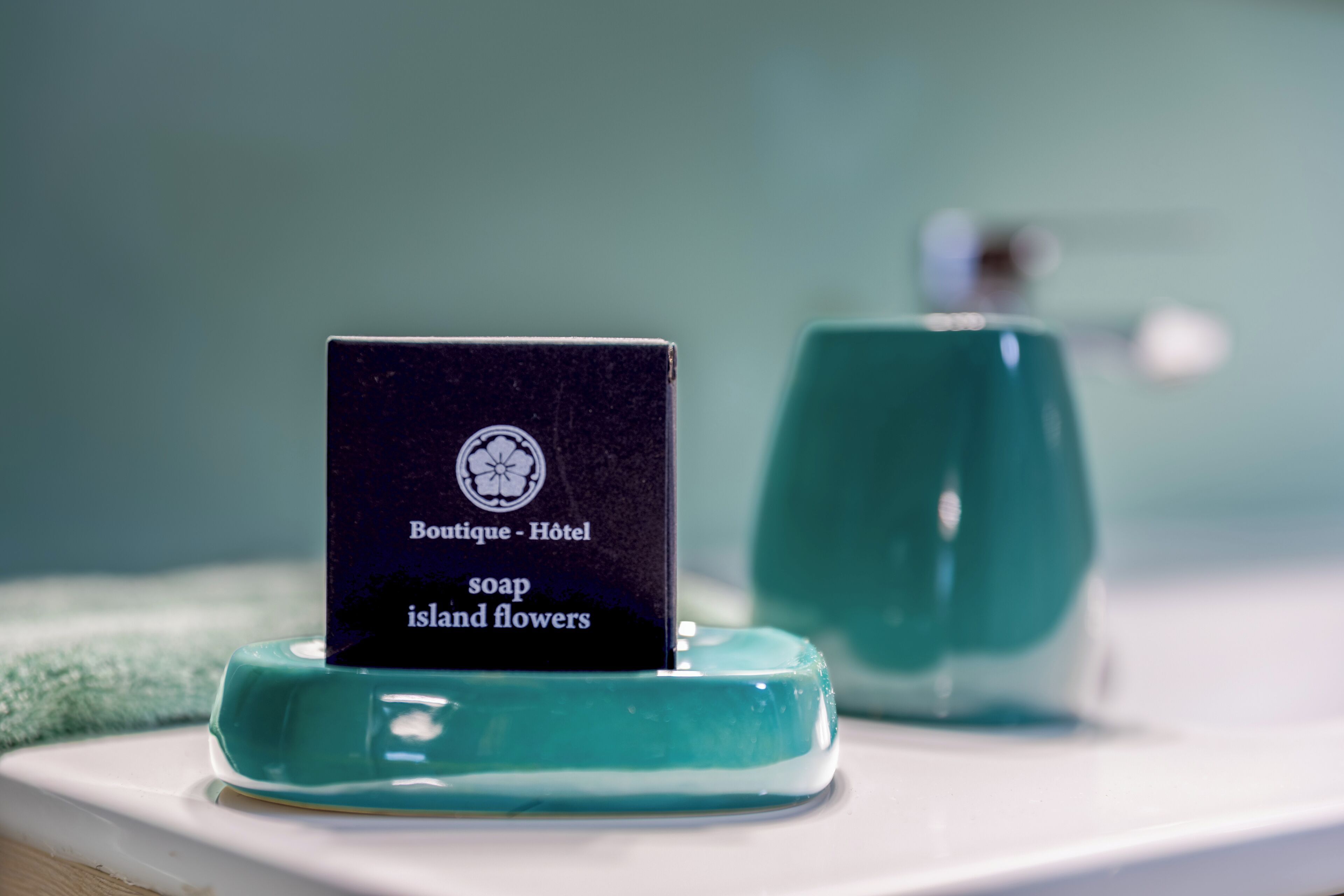 Una pastilla de jabón con la etiqueta 'Boutique - Jabón de hotel flores de la isla' sobre un plato de jabón color turquesa, con un dispensador de jabón color turquesa al fondo.