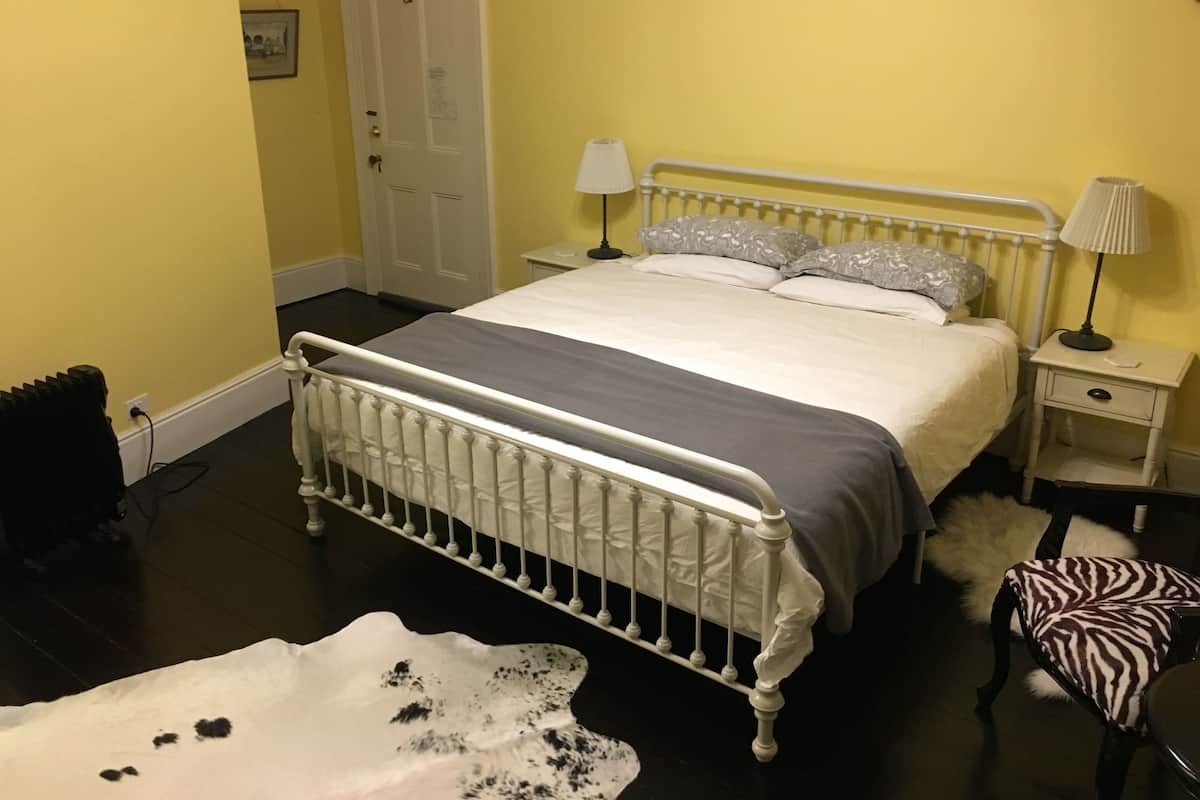 Quarto com estrutura de cama de metal branco, colcha cinza, duas luminárias de cabeceira, porta branca e uma pequena cadeira branca com almofada estampada com zebras.