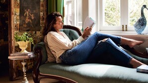 A person reading a book on a green sofa in a room with a large window, a wall with a collage of pictures, and a small table with a vase and books. - Boscobel of Tasmania (West Ulverstone)