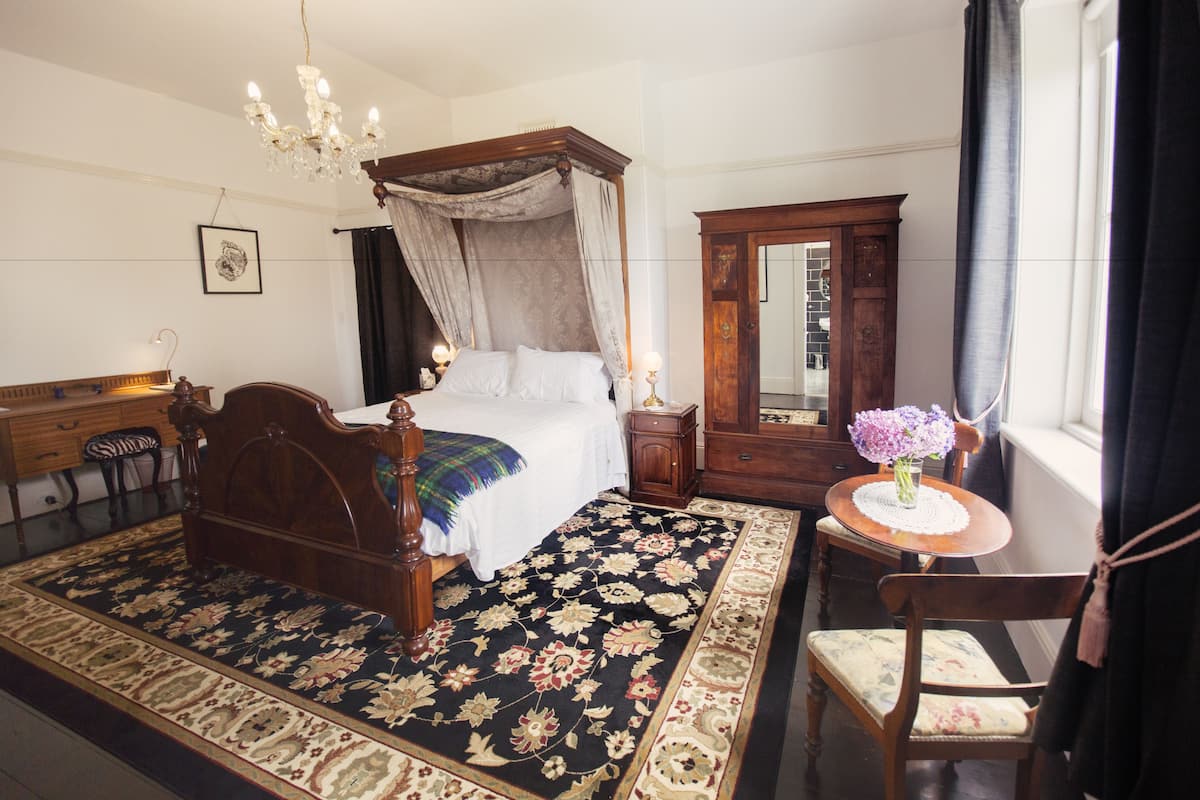 Quarto com cama de dossel, mesa de cabeceira de madeira, pequena mesa com um vaso de flores, uma cadeira e um espelho grande.