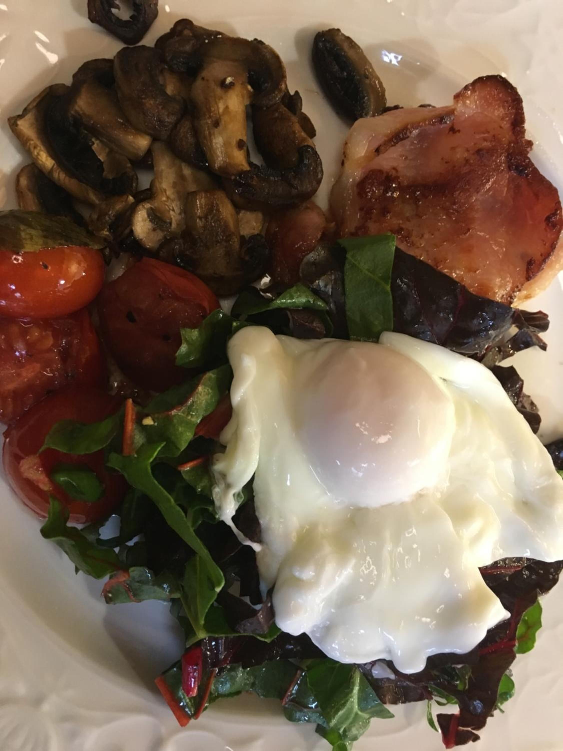 A plate of food with a poached egg, mushrooms, bacon, tomatoes, and greens.