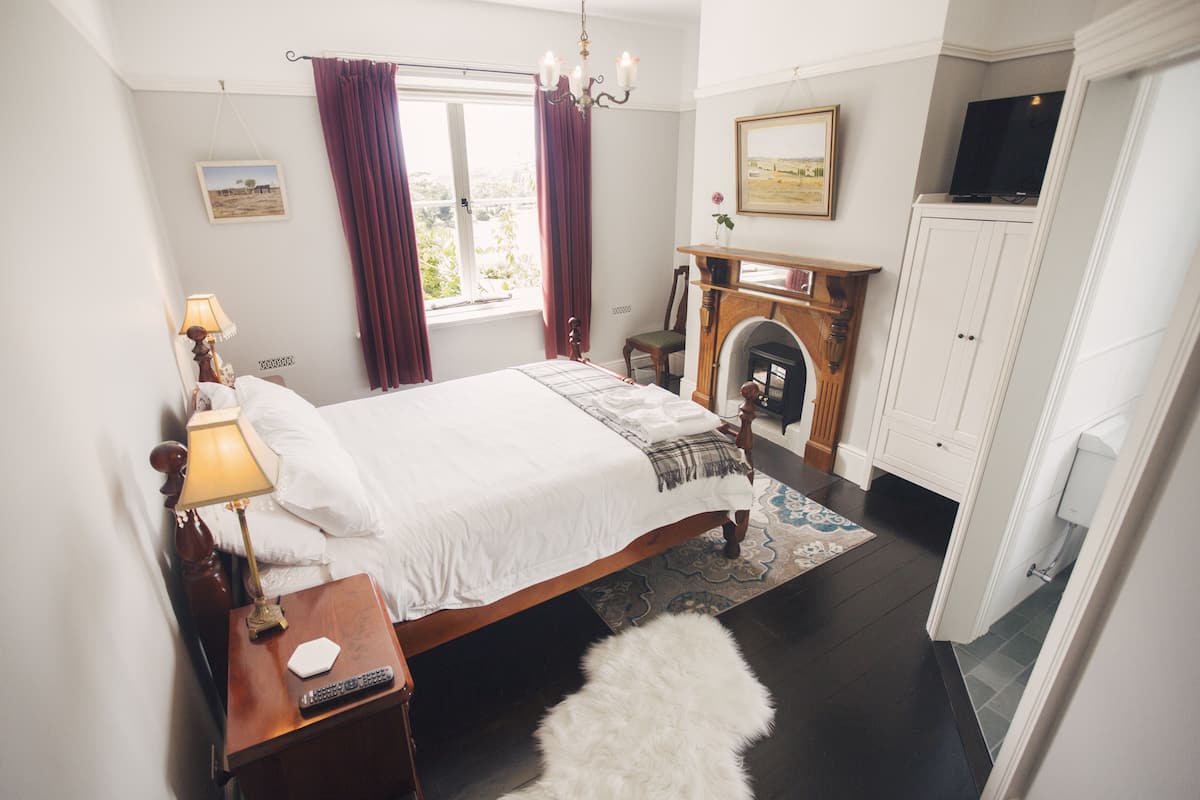 Quarto com cama, mesa de cabeceira com abajur, lareira, guarda-roupa e janela com cortinas.