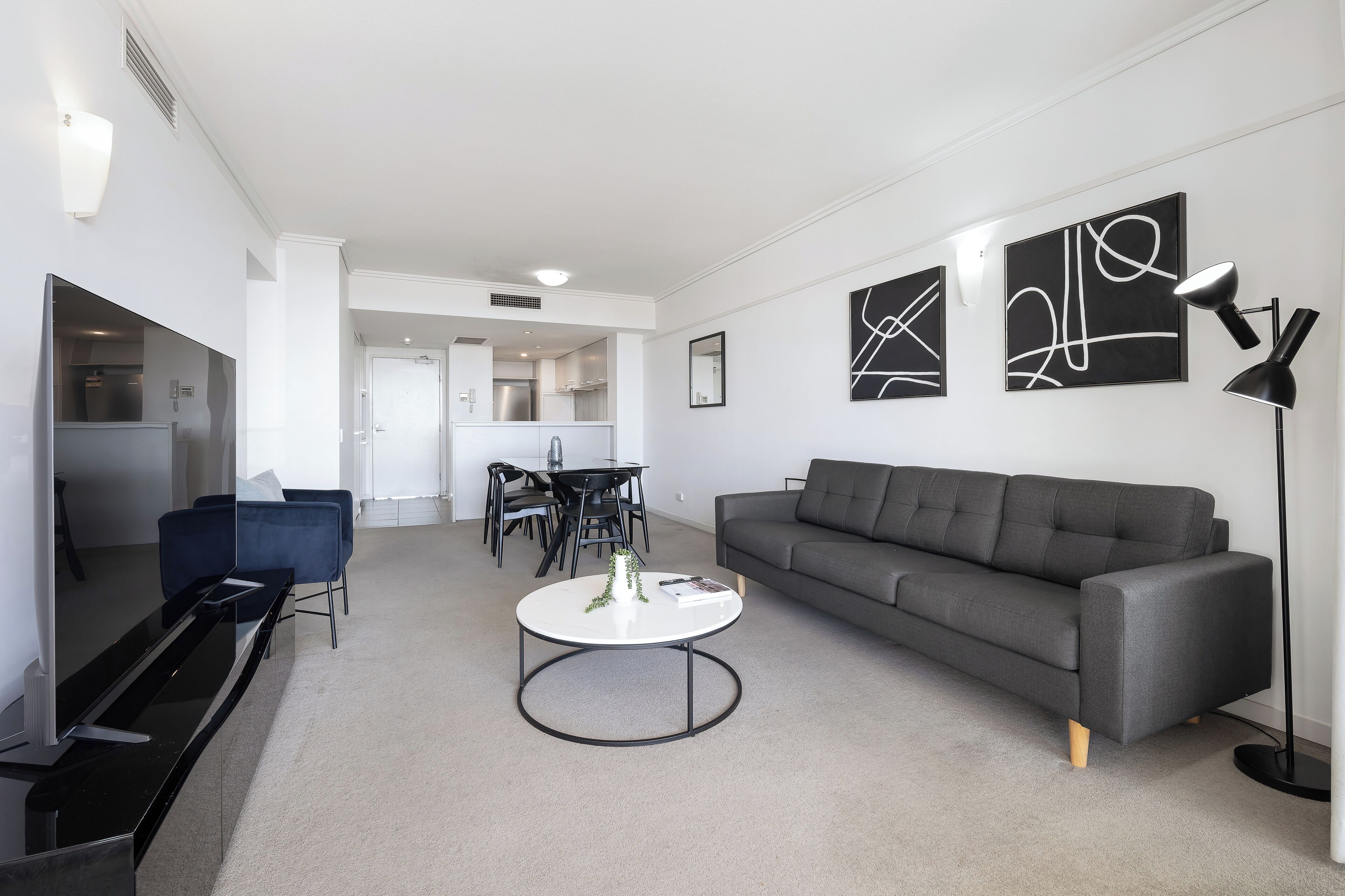 A modern living room with a grey sofa, a round coffee table, and a TV on a stand. There is a dining area with a table and chairs in the background.