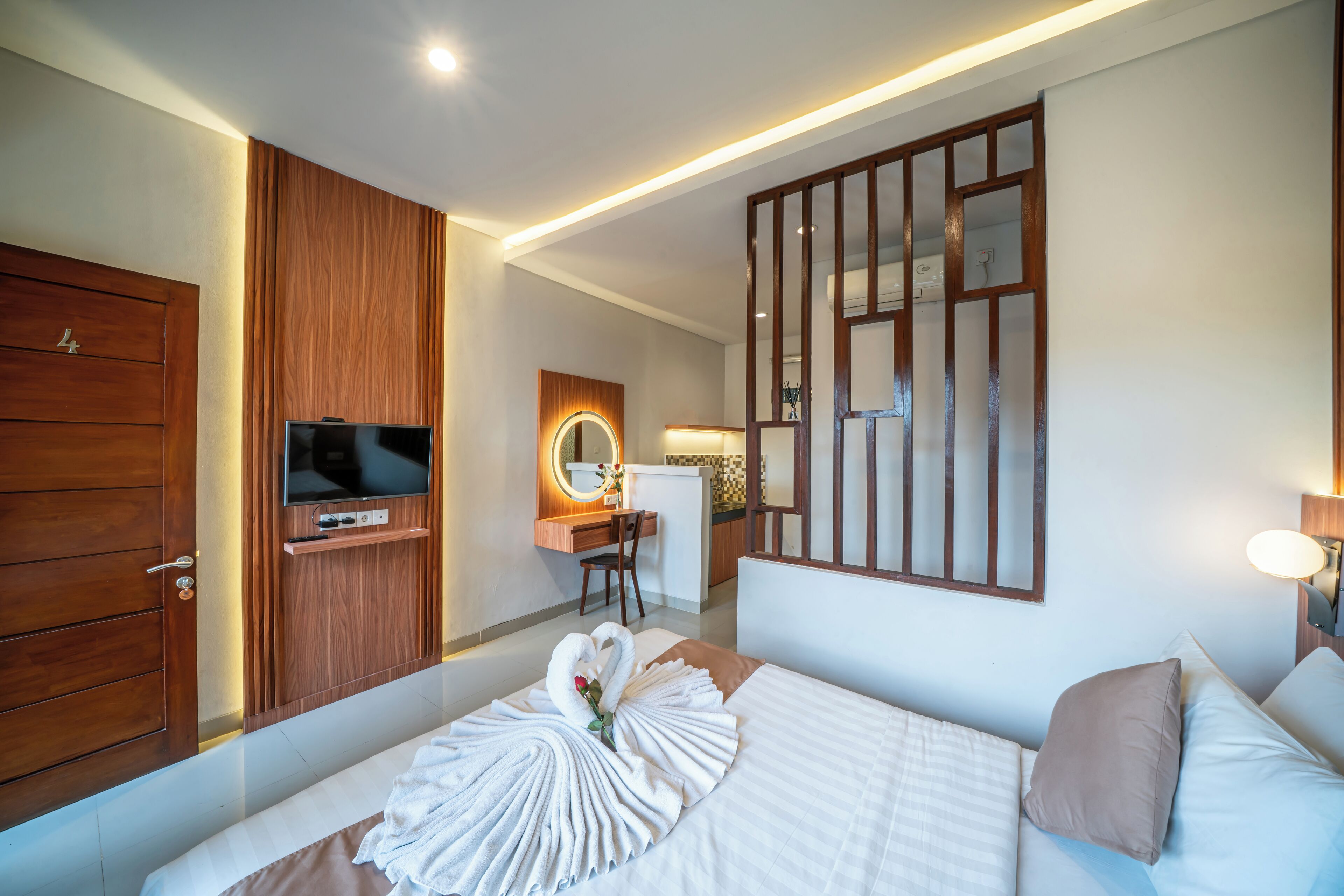 Habitación de hotel con armario de madera, televisor, escritorio con silla, cama con toallas y una rosa, y un espejo grande.