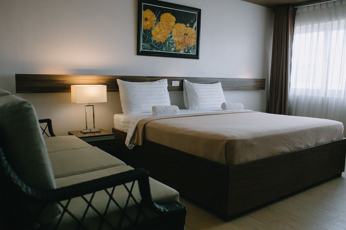 Una habitación de hotel con una cama grande, una mesita de noche con lámpara, una silla y un cuadro de flores en la pared.
