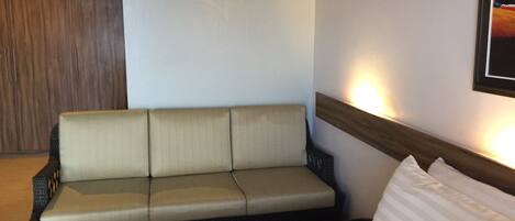 A hotel room with a beige sofa, a bed with white pillows, and a wall-mounted light fixture.