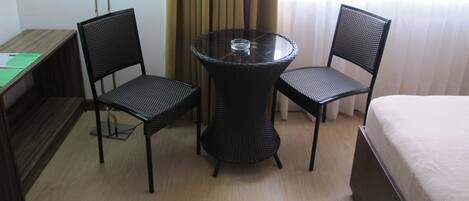 Habitación de hotel con una mesa redonda de vidrio, dos sillas de mimbre, un escritorio con lámpara y una cama.