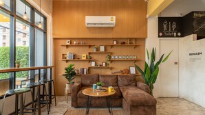 A cozy lounge area with a brown sofa, a small round table, and a bar stool. Shelves with books and decorative items are visible. - BEDGASM Hotel x Café at Nimman (Chiang Mai)