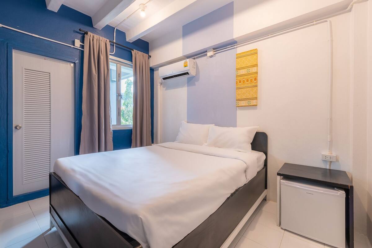 A bedroom with a bed, a bedside table, a small refrigerator, and a window with curtains.