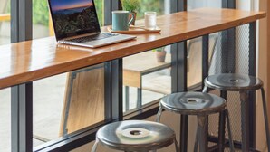 A workspace with a laptop, a mug, and a plant on a wooden table by large windows with hanging light bulbs. - BEDGASM Hotel x Café at Nimman (Chiang Mai)