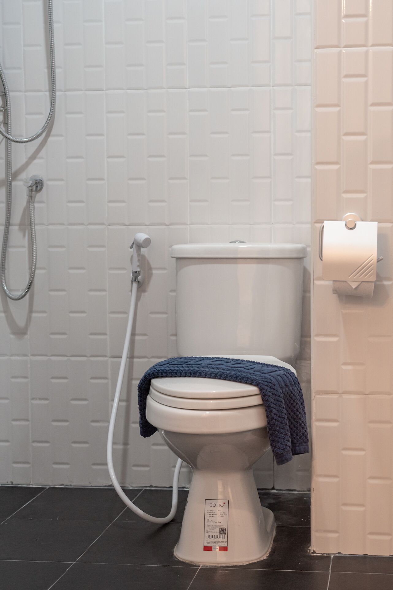 A white toilet with a blue towel, a shower hose, and a toilet paper holder on a white tiled wall.