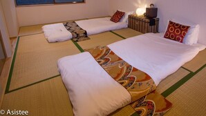 Een traditionele Japanse kamer met tatami matten, twee bedden met witte beddengoed, een kleine tafel met een lamp, en kunstwerken aan de muren.