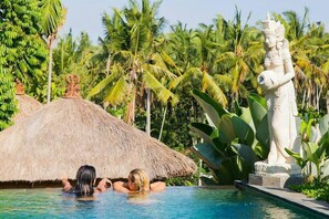 Une piscine où deux personnes se détendent sous un toit de chaume, entourées de palmiers et d’une statue.