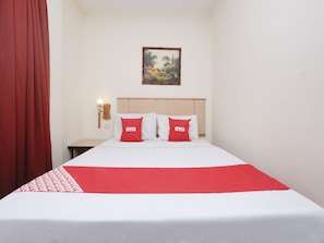 Une chambre d’hôtel avec un lit, deux oreillers rouges portant la marque « OYO », une tête de lit en bois, une table de chevet avec une lampe et un tableau accroché au mur.