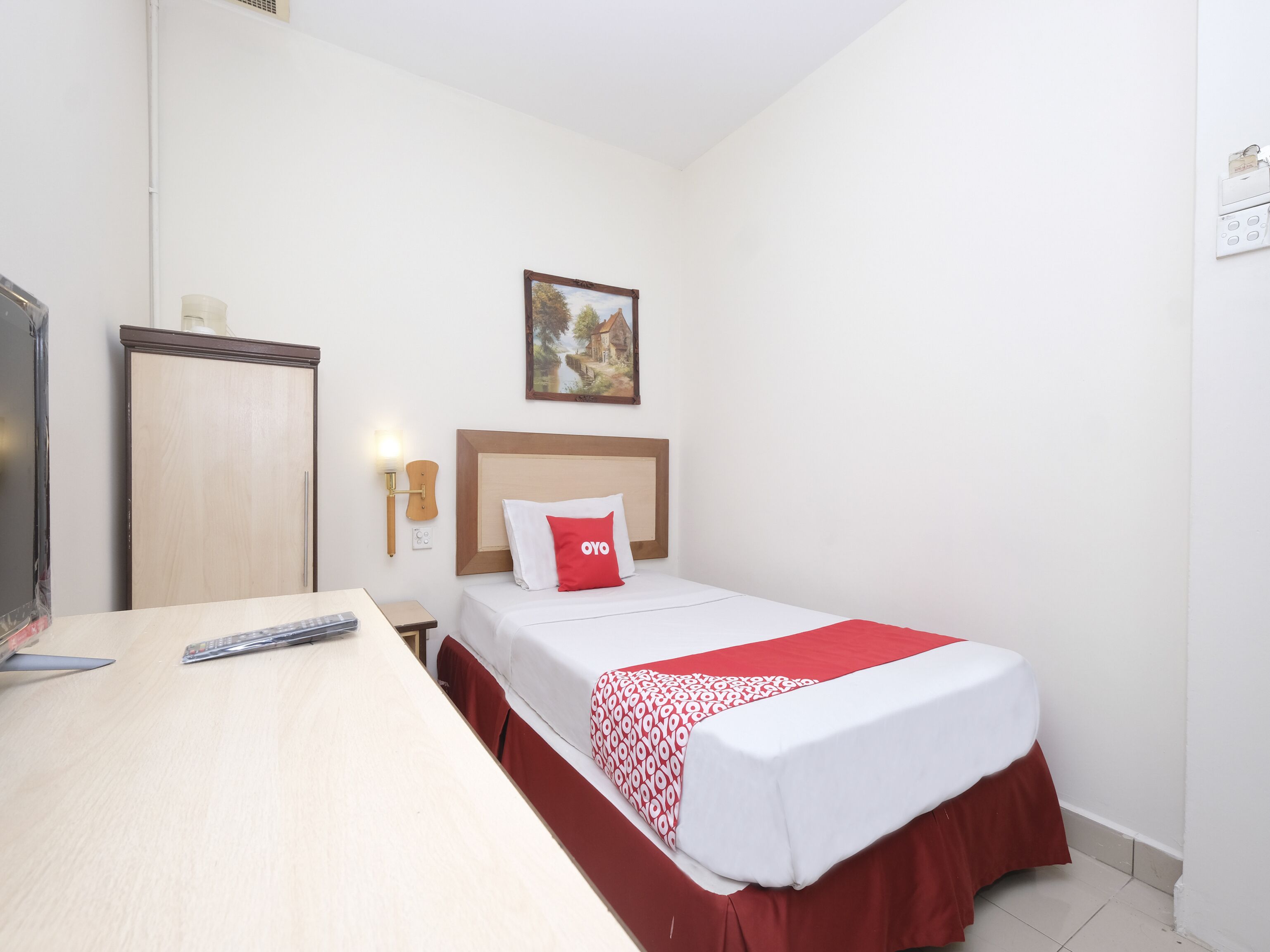 Una habitación de hotel con una cama individual, una mesita de noche de madera, un escritorio con un control remoto y una lámpara de pared.