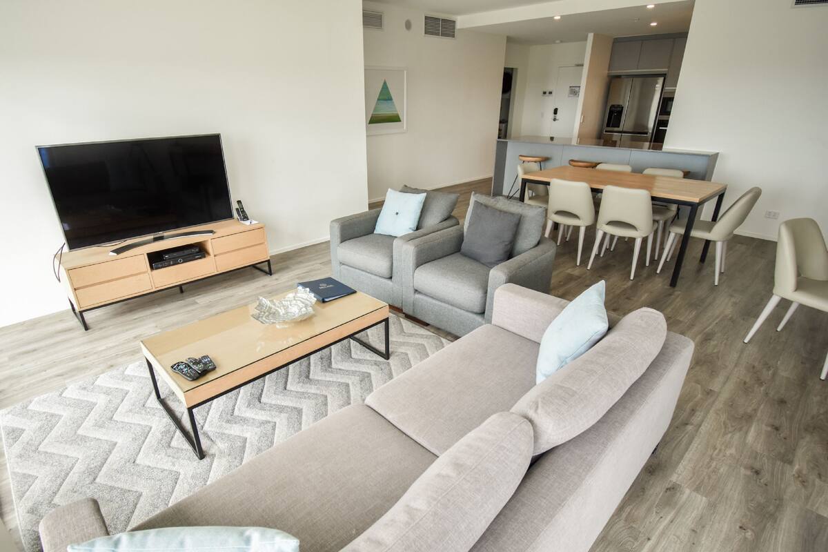 A modern living room with a large flat-screen TV, a sofa, armchairs, a coffee table, and a dining area with chairs and a table.