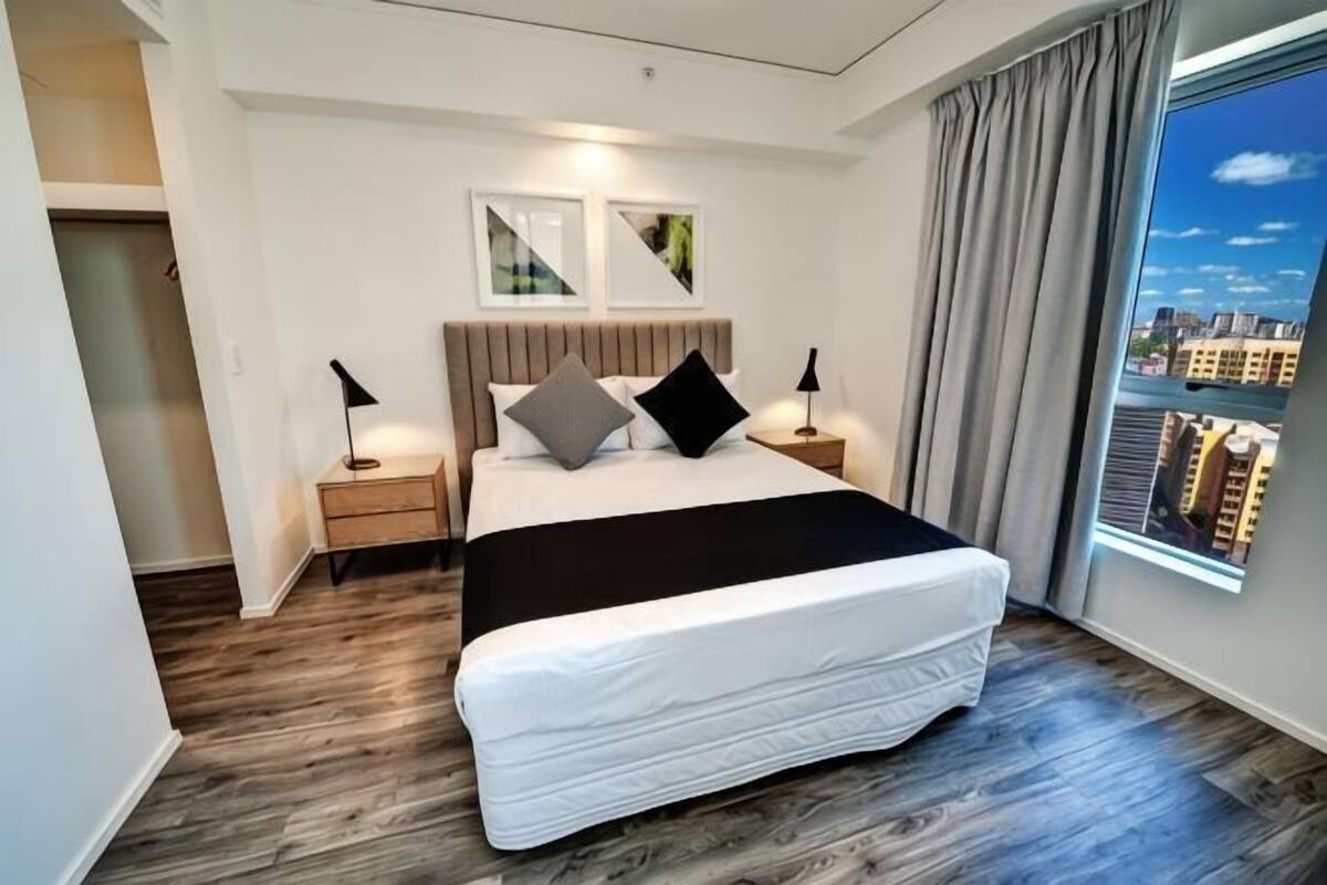 A modern bedroom with a large bed, bedside lamps, a city view, and a wooden floor.