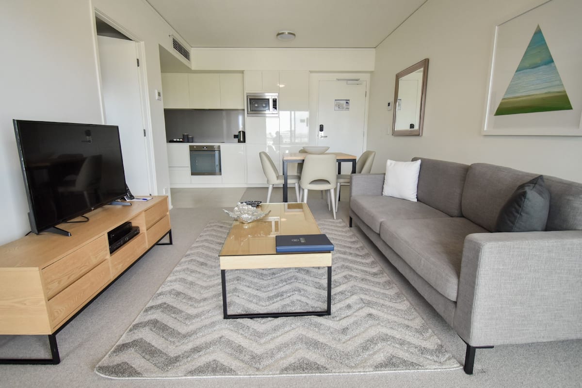 A modern living room with a flat-screen TV, a wooden coffee table, a grey sofa, and a kitchen in the background.