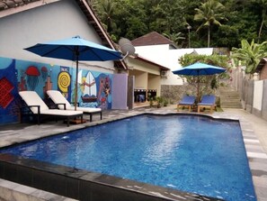 Una piscina con sillas de descanso y sombrillas, rodeada por un mural y una escalera que lleva a una casa.