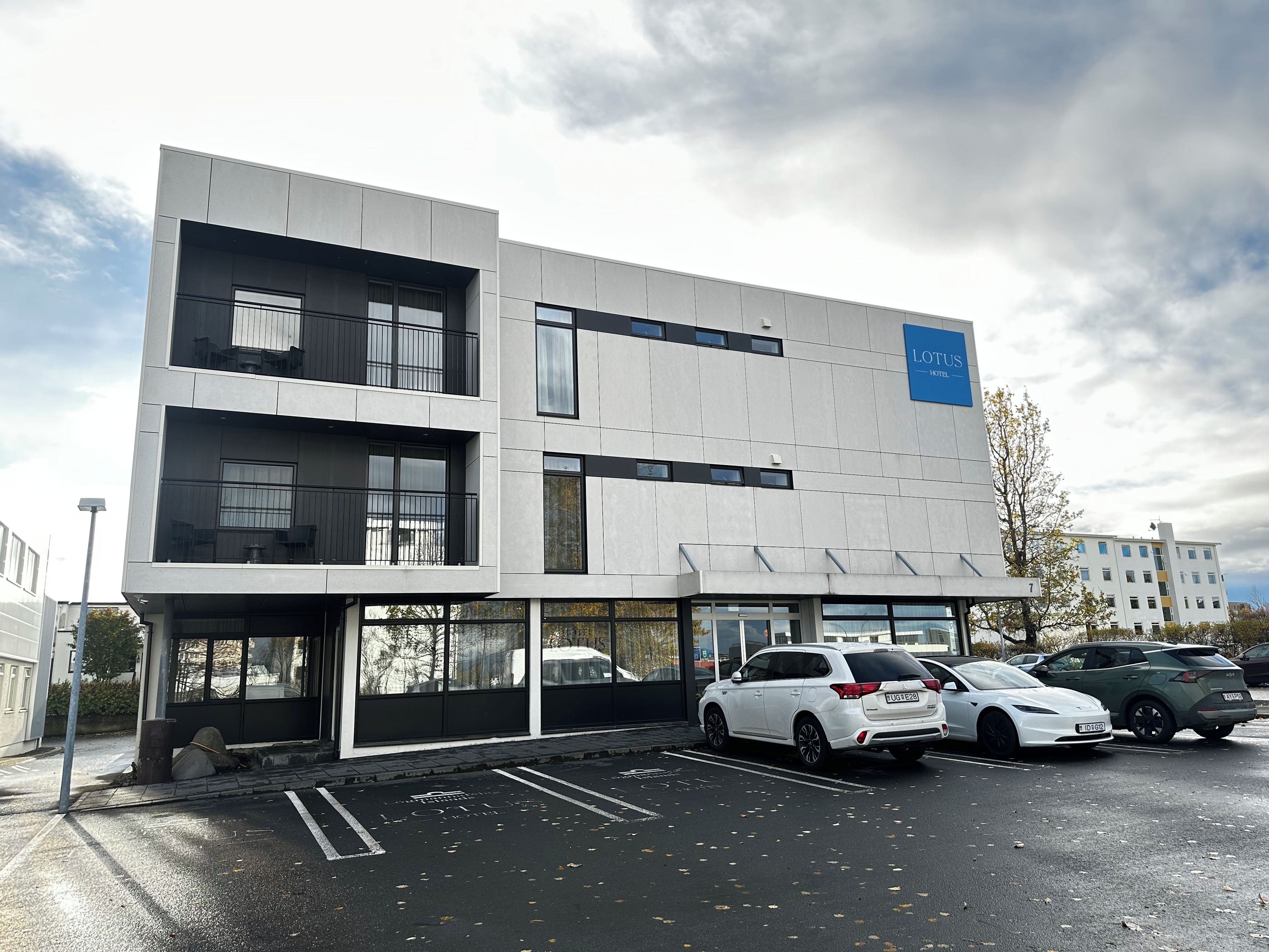 Un bâtiment moderne, avec une enseigne « LOTUS HOTEL » sur le côté, un parking avec plusieurs voitures et un ciel dégagé.