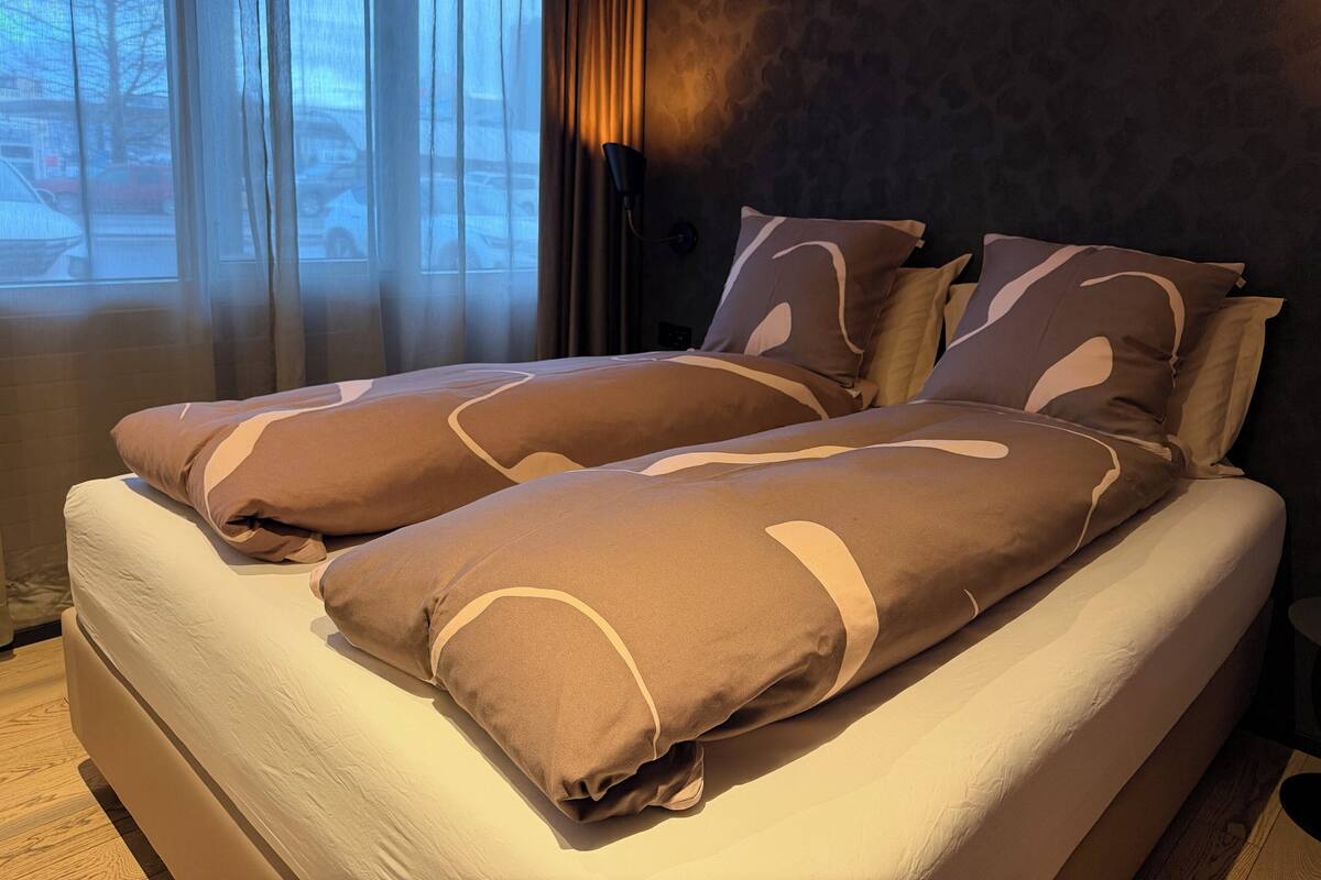Una cama con colchón blanco y almohadas marrones, una ventana con cortinas traslúcidas y una pared oscura con textura.