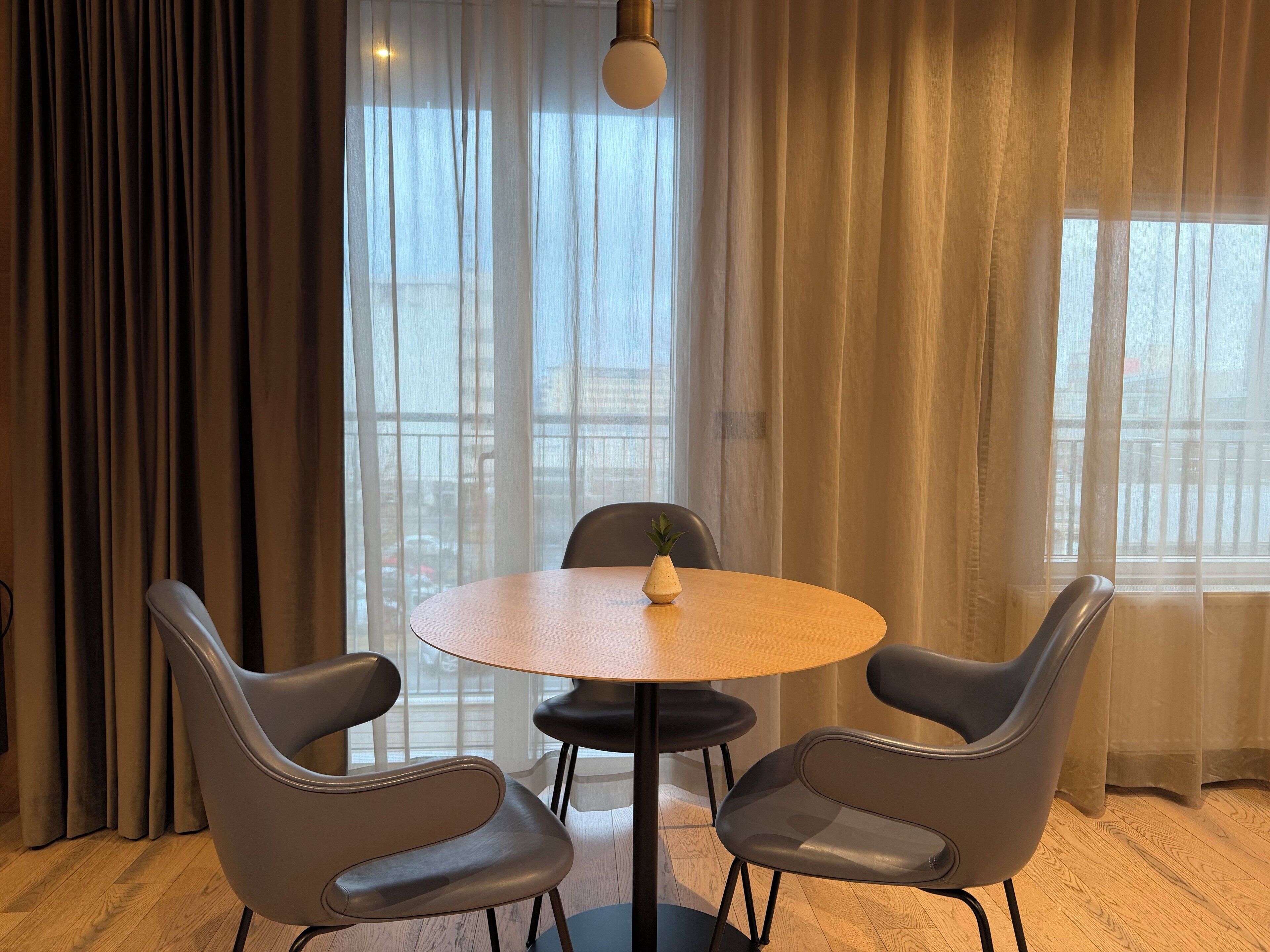 Una habitación con una mesa redonda, dos sillas, una planta en maceta y ventanas amplias con cortinas traslúcidas.
