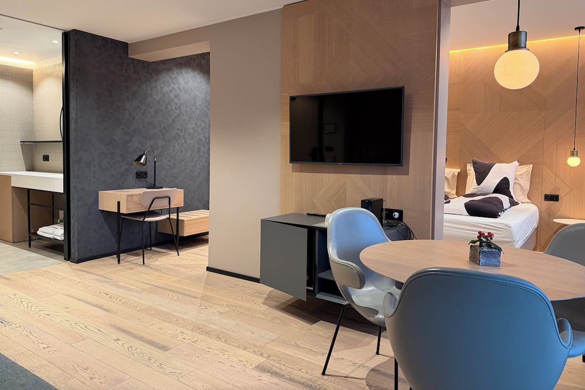 A modern hotel room with a wooden floor, a dining area with blue chairs, a flat-screen TV mounted on the wall, and a bed with pillows and a blanket.