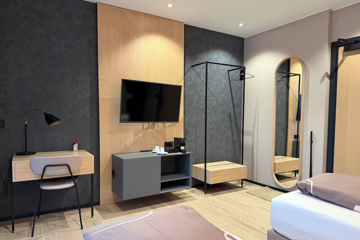 Una habitación de hotel moderna con escritorio, silla, cama, televisor, armario y un espejo grande.