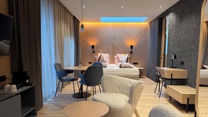 Una habitación de hotel moderna con cama, escritorio, silla y sofá. Hay un televisor a la izquierda y una ventana con cortinas.