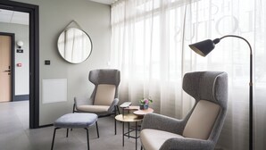 Una habitación de hotel moderna con dos sillones tapizados en gris, una mesita redonda con lámpara y un ventanal con cortinas de gasa.