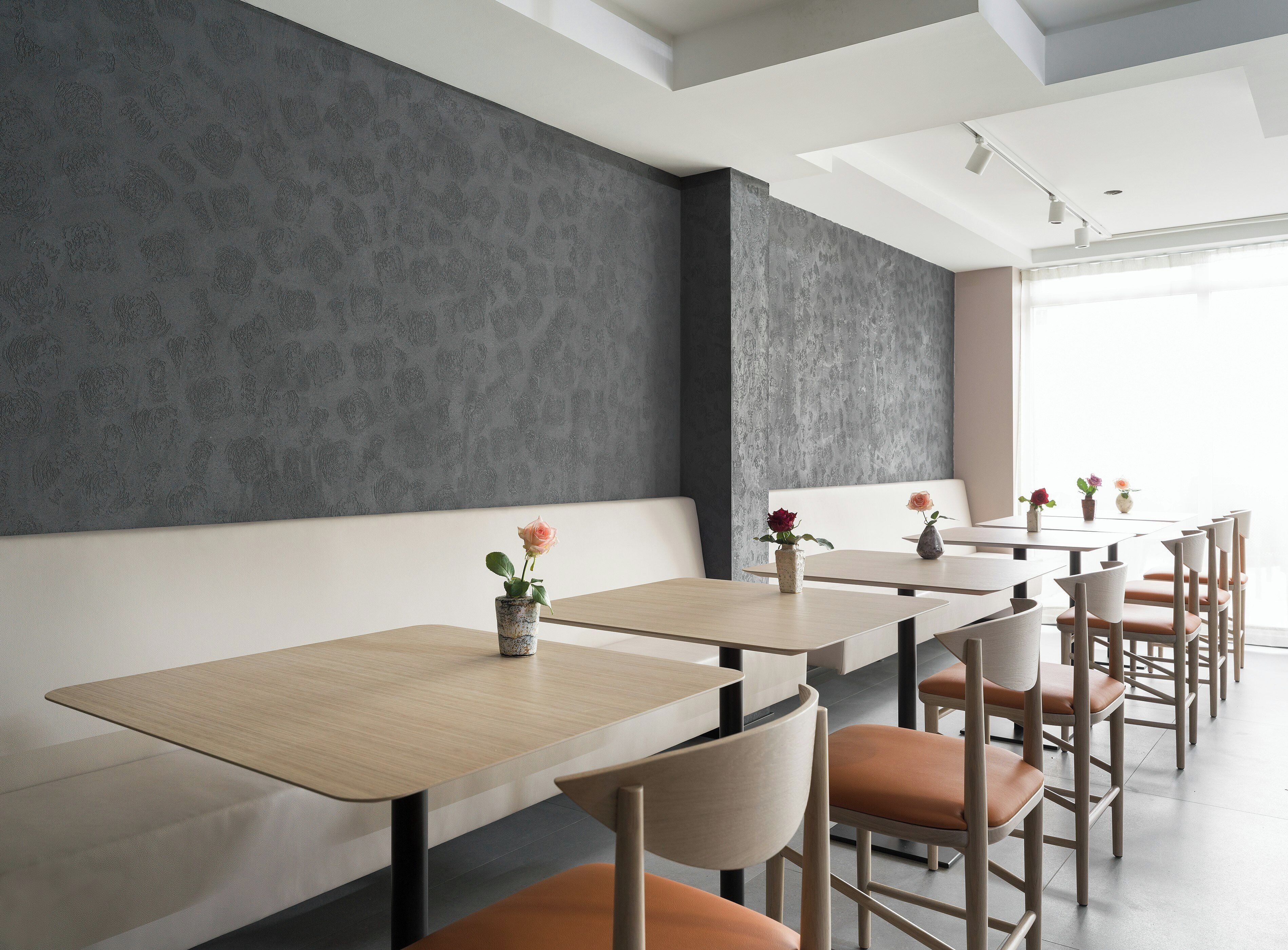 Uma sala de jantar moderna com mesas e cadeiras compridas, parede texturizada cinza e janela que oferece luz natural.