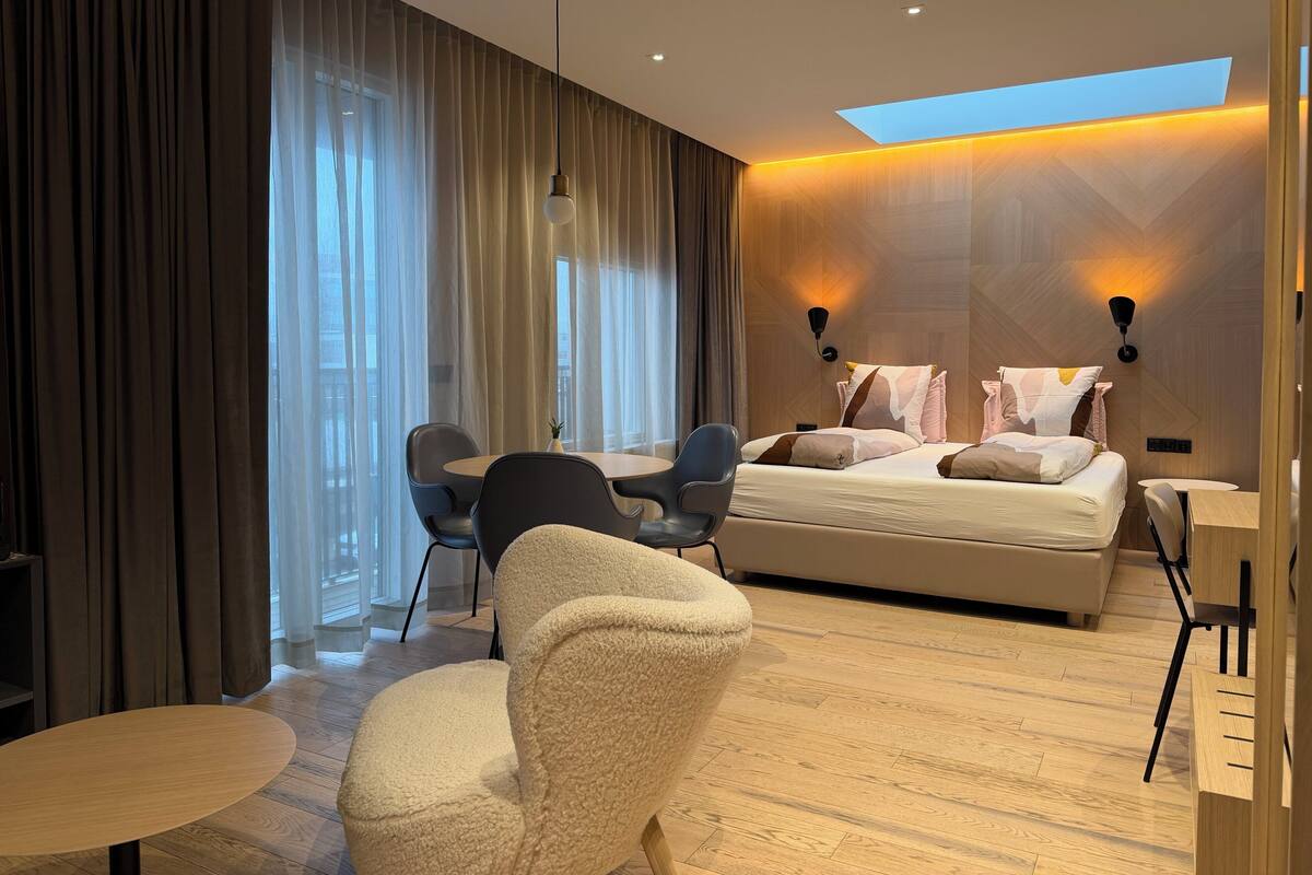 Habitación de hotel moderna con cama, escritorio y silla. Cuenta con un ventanal con cortinas, una lámpara de techo y una lámpara de pared.