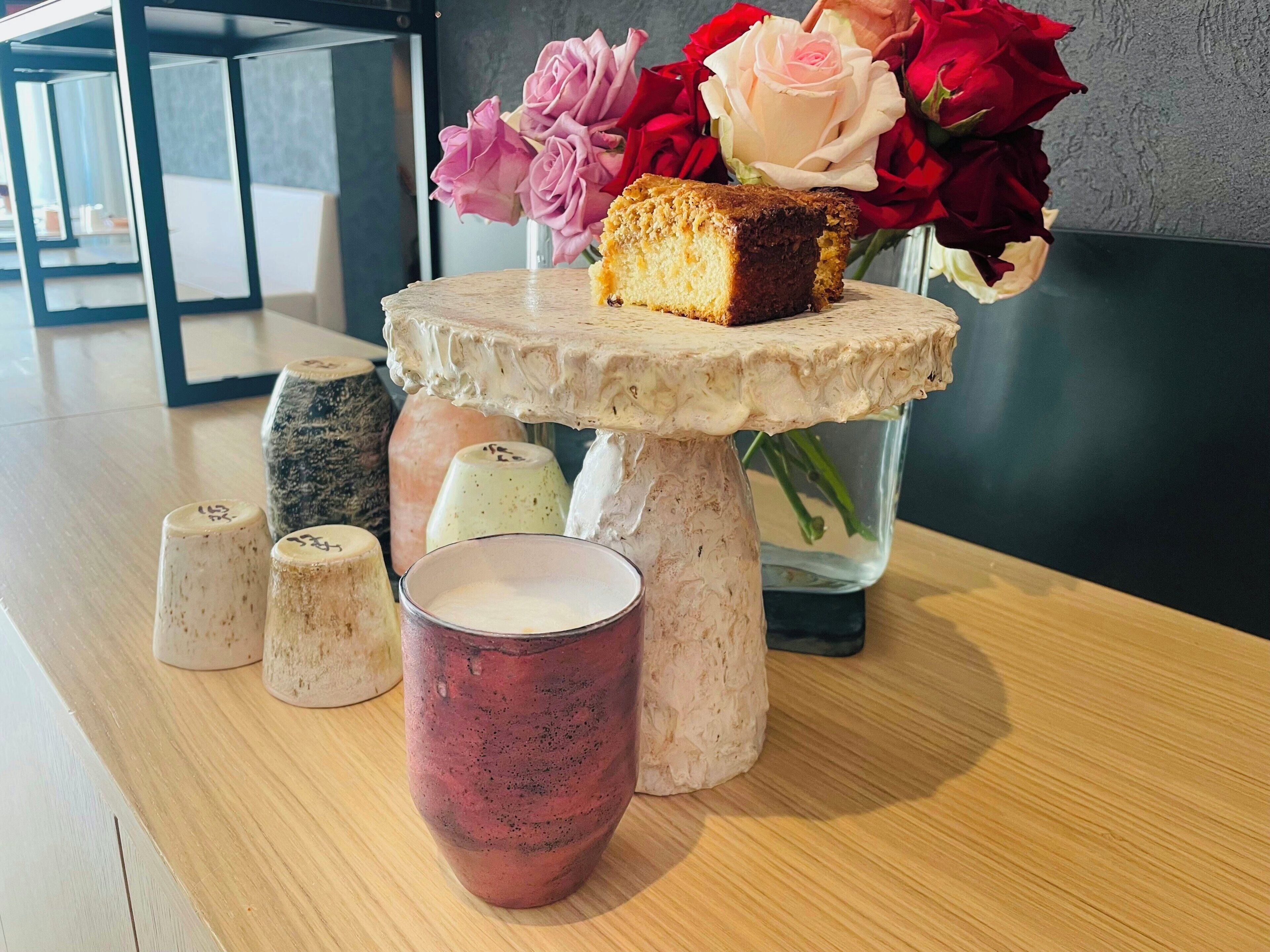 A table with a cake on a pedestal, a glass of milk, and a vase with flowers.