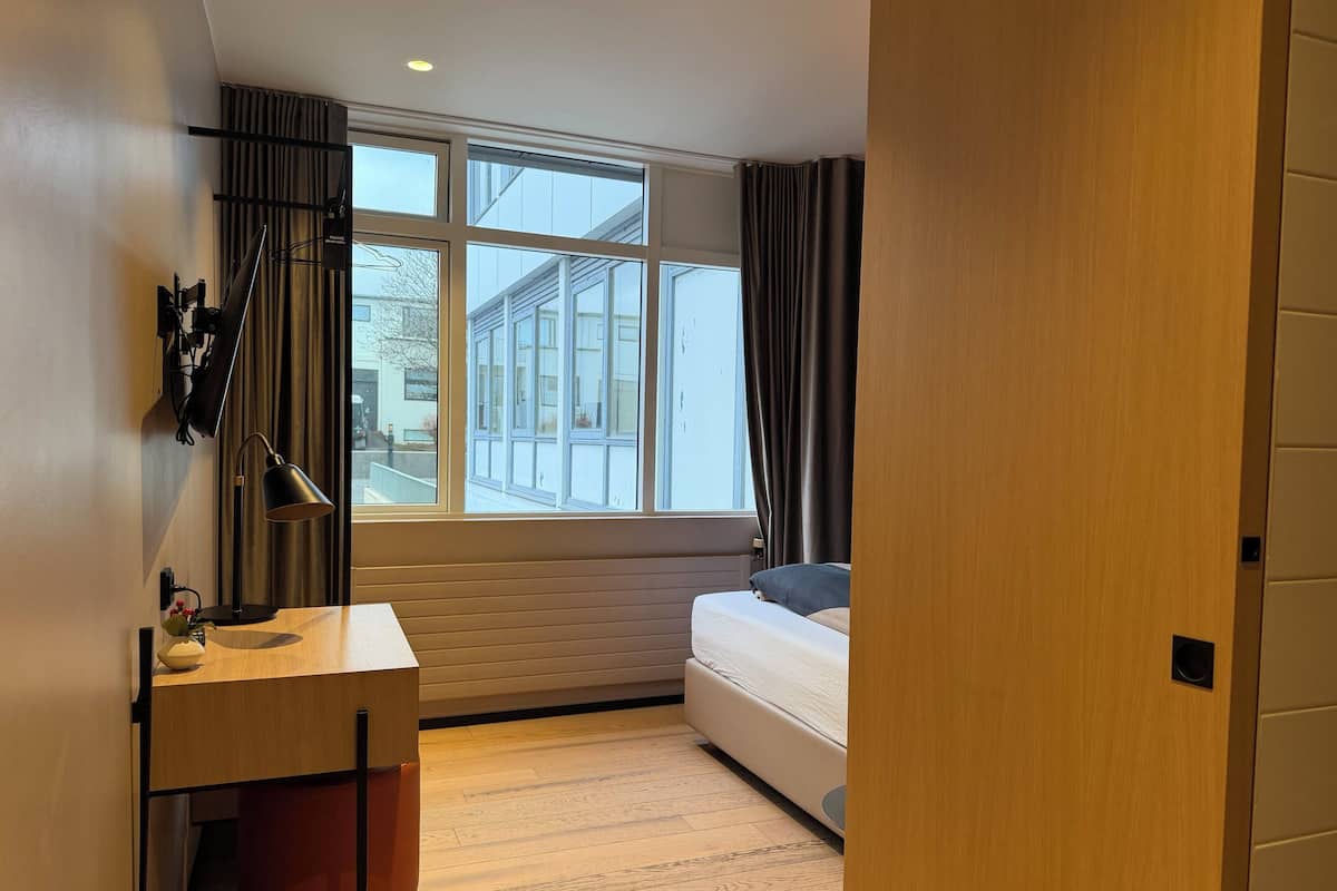 Un dormitorio con una cama, un escritorio con lámpara, una ventana y vista a un edificio.