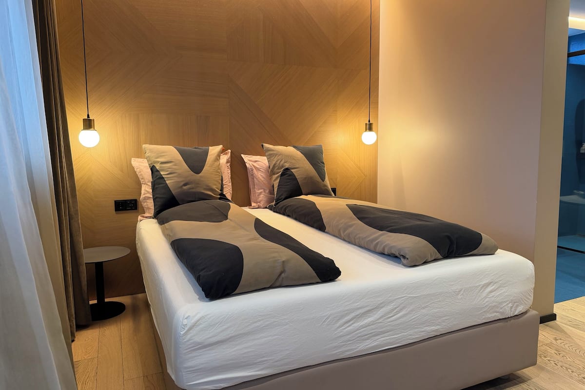 Un dormitorio moderno con cabecera de madera, cama con ropa de cama blanca y almohadas estampadas, y dos lámparas colgantes.