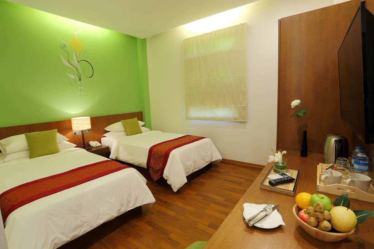 A hotel room with two beds, a wooden desk with a bowl of fruit, a coffee maker, and a window with blinds.