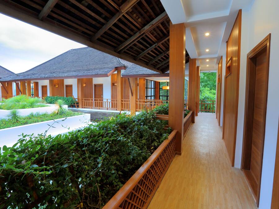 A corridor with wooden railings, a covered area with a view of a garden, and a pathway leading to a building entrance.
