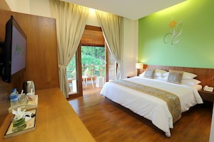 A hotel room with a wooden floor, a bed with white bedding, a nightstand with a phone, a desk with a water bottle, a coffee maker, and a small table with a flower vase.