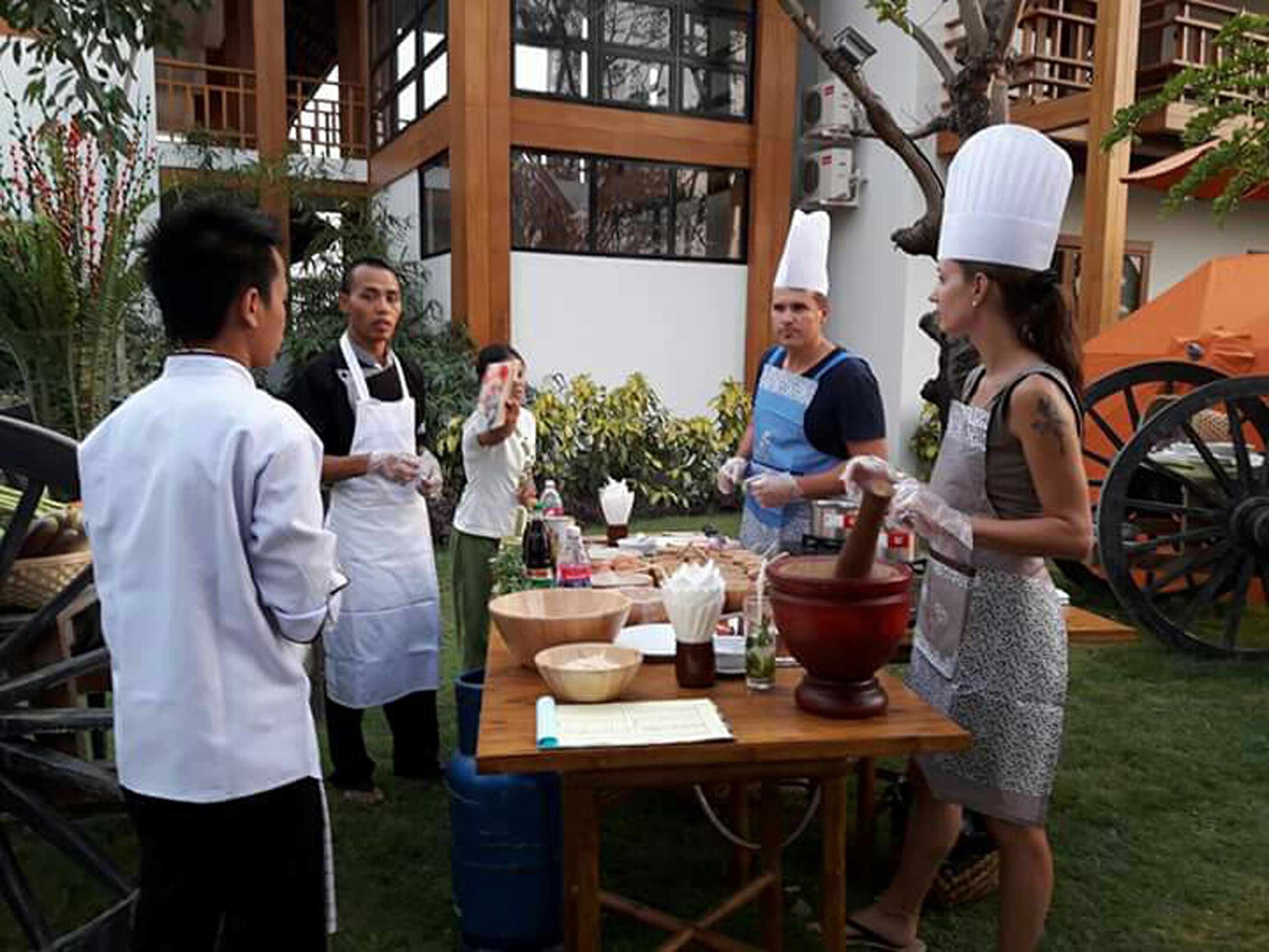 A group of people in a courtyard with a wooden table, a large bowl, and a mortar.