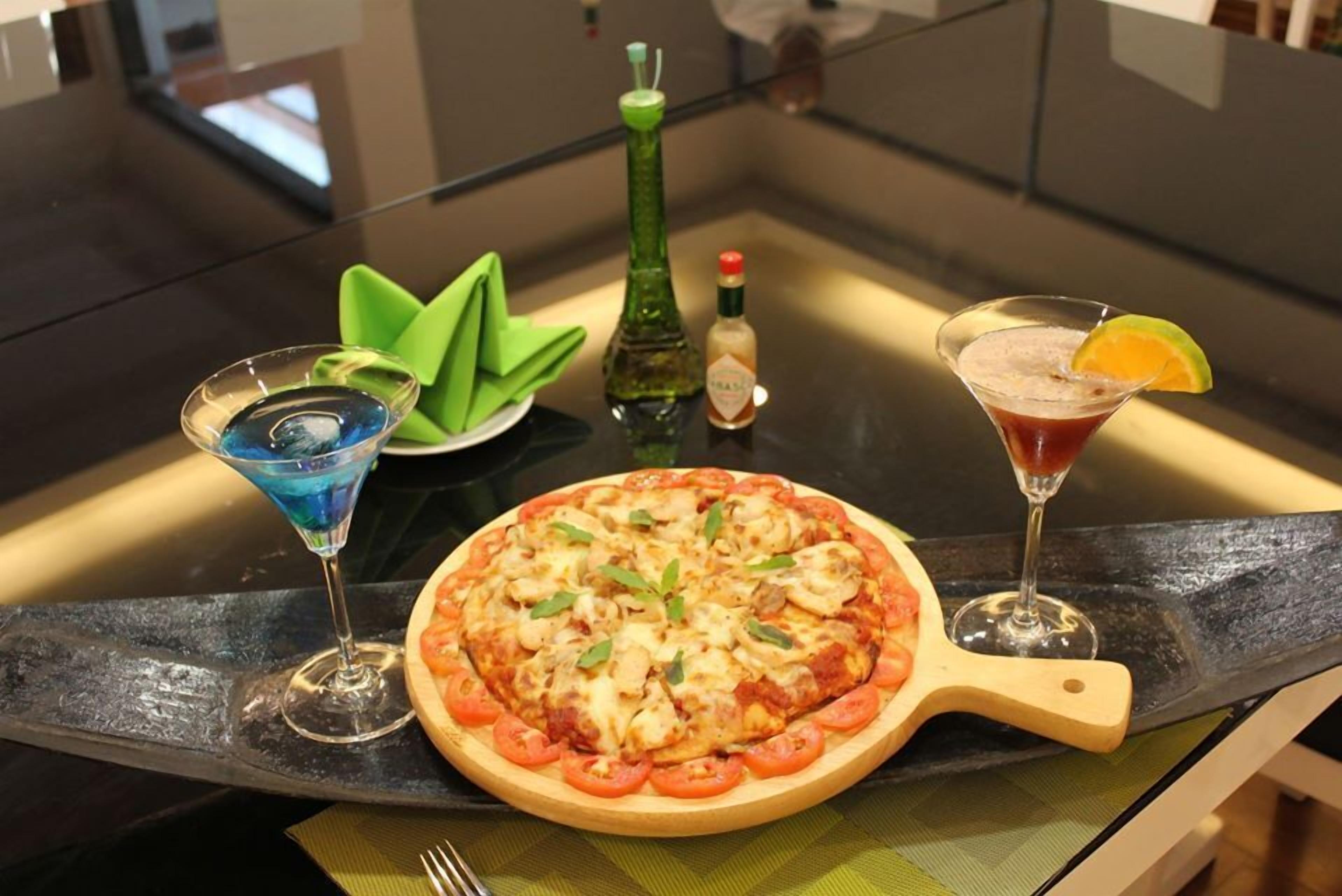 A pizza on a wooden board with a green leaf decoration, a martini glass with an orange slice, and a bottle of sauce on a kitchen counter.