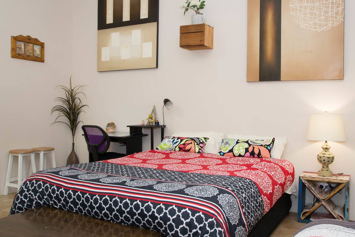 A bedroom with a large bed, a desk, a chair, a small table, and a lamp.