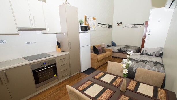 A compact kitchen with a microwave, oven, and a refrigerator. There is a dining area with a table and chairs, and a sofa in the background. - Westside Studio Apartments (Armidale)