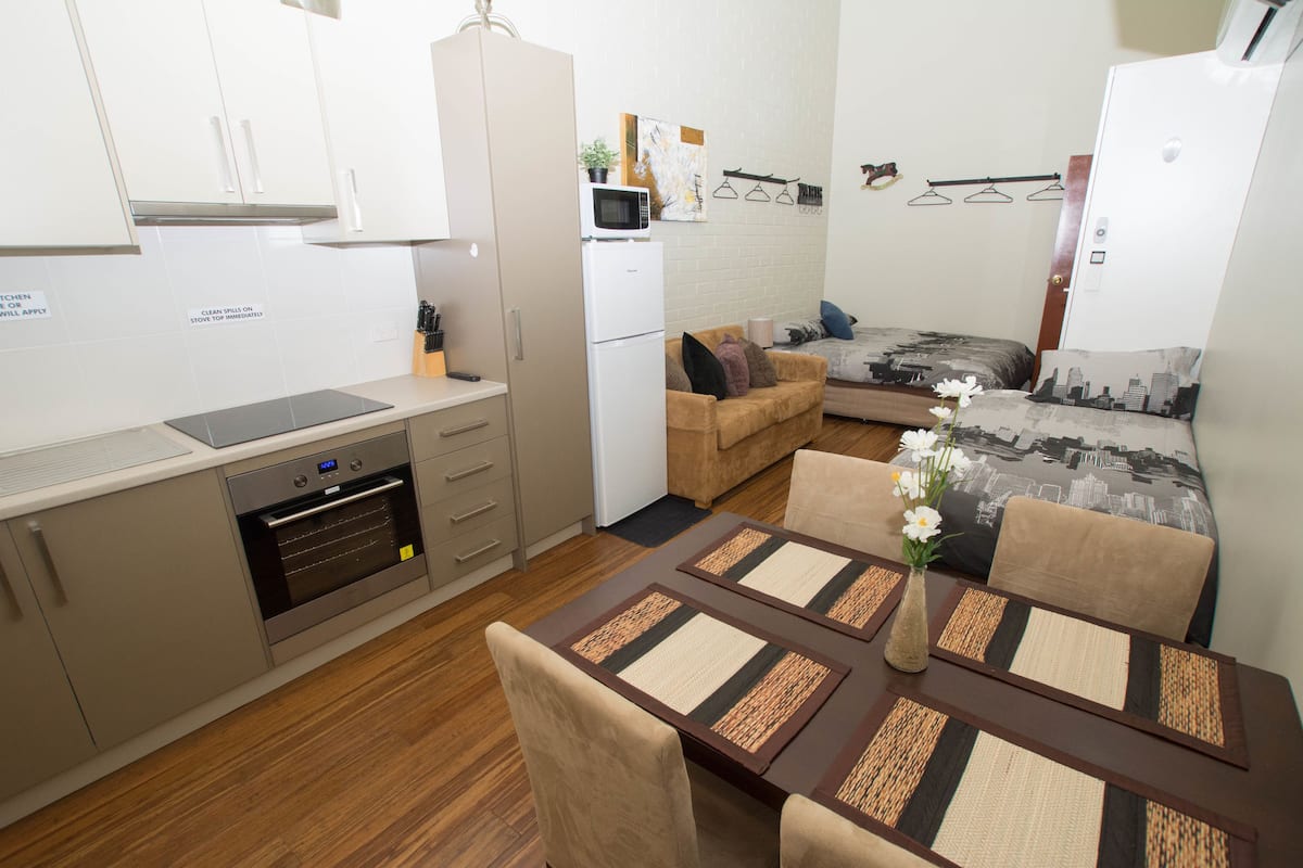 A compact kitchen with a microwave, oven, and a refrigerator. There is a dining area with a table and chairs, and a sofa in the background.