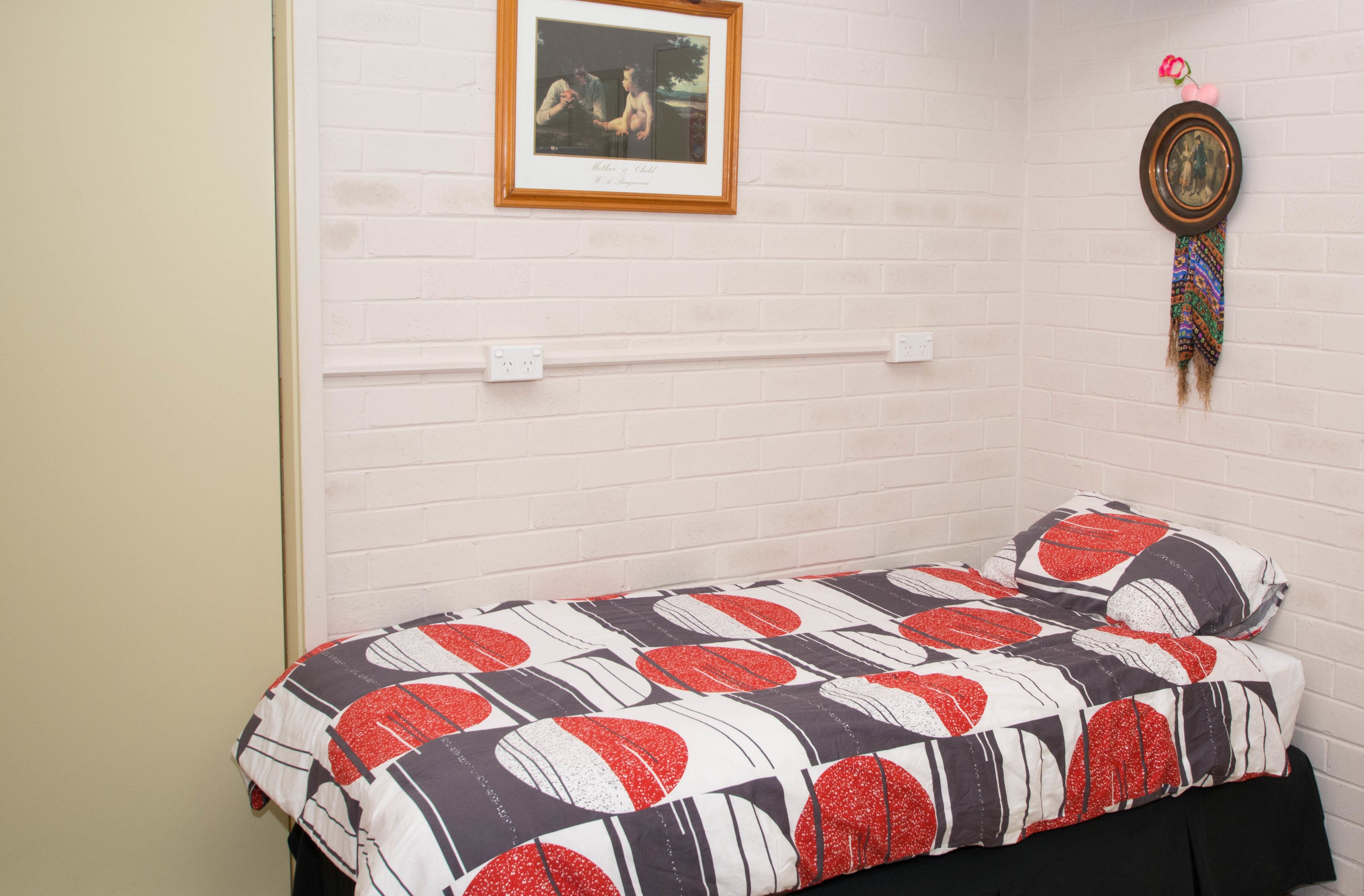 Una habitación pequeña con cama individual, pared de ladrillo blanco, un cuadro enmarcado y un adorno colgante.